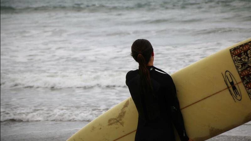 woman in wetsuit, holding surfboard and looking towards water