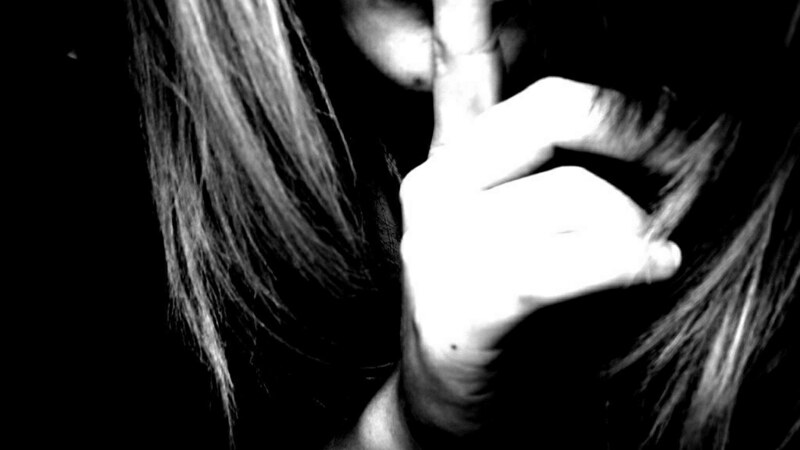 grainy black and white photo of woman&#39;s mouth and chin, with finger to her lips, mouthing &quot;shh!&quot;