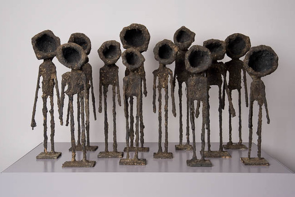 photo of sculpture, composed of twelve small sculptures of human-like figures with hollowed out cylinders in place of heads