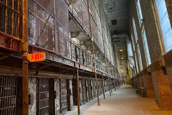 An empty prison hallway, with two stories of rusty cells on one side and windows on the other.