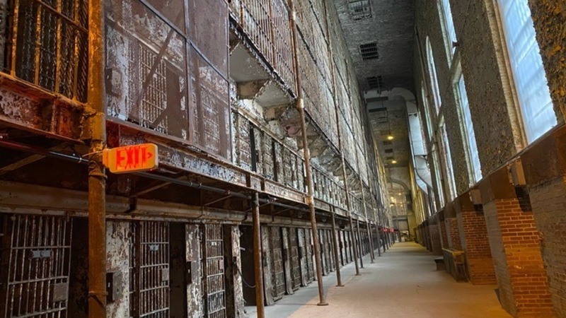 An empty prison hallway, with two stories of rusty cells on one side and windows on the other.