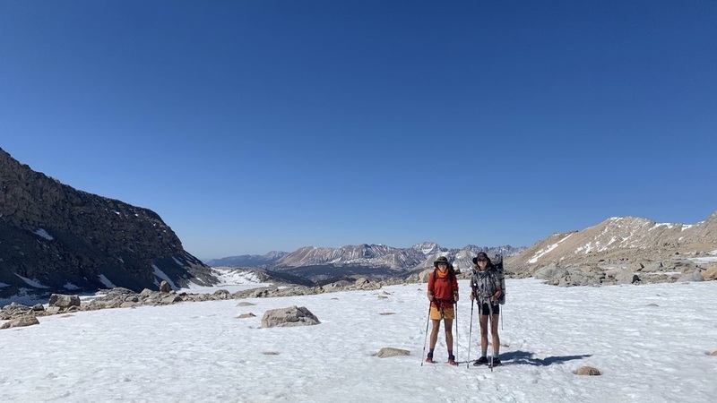 Two young women stand on snowy ground in hiking gear, against a mountainous backdrop and bright blue sky.