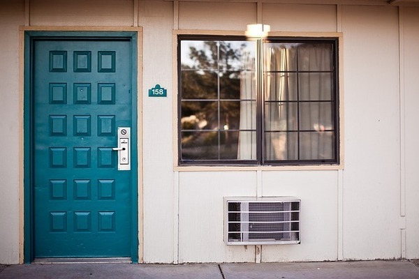 Photograph of the outside of a hotel room with a blue door, AC unit, and window with half open white curtains