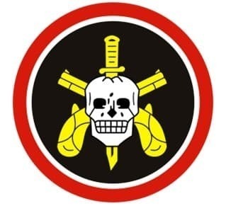 a logo consisting of a red circle with a black circle inside it. Inside the black circle is a white skull in front of two crossed yellow guns with a yellow knife inserted in the skull from top to bottom.
