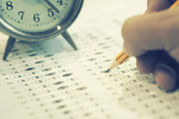 Hand holding a pencil hovering over a scantron for multiple choice tests with part of an alarm clock in the background