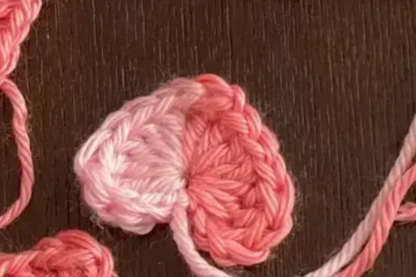 Crocheted hearts in various shades of pink and light pink yarn strands lie on a dark surface. One heart is two-toned.