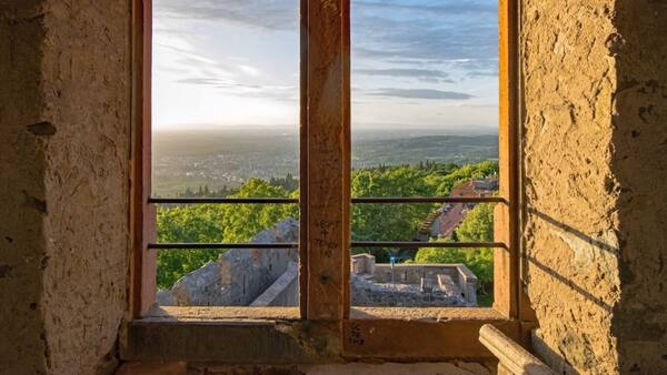 View through open rectangular double windows set in a rough stone wall. Tourist graffiti in black ink is faintly visible on the verticle beam separating the apertures. Golden sunlight bathes the stone wall in the foreground as well as green trees and a distant town far below, and thin clouds hover in a bright blue and white sky above.