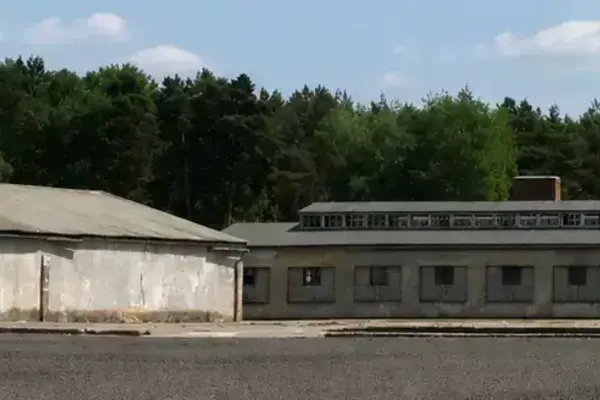 Two long, grey barracks buildings in a gravel yard, backed by a dense green forest under a partly cloudy sky.